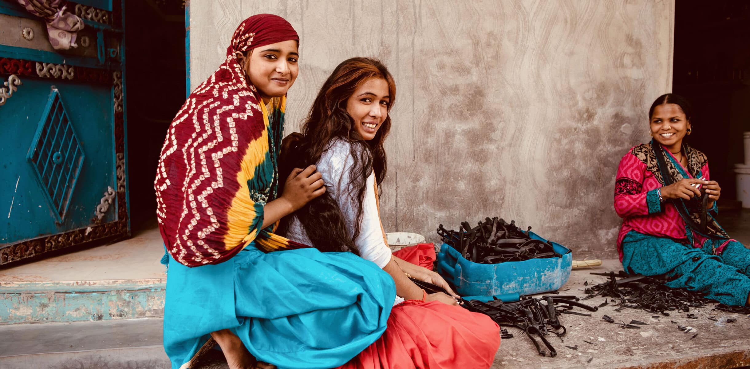 Three informal workers in Delhi, India, sit outside a building, smiling and assembling plastic components as part of their work, 2018.