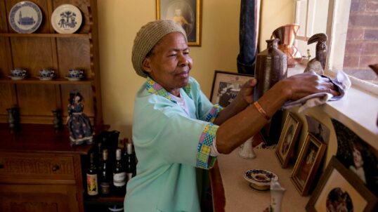 Anna Nkobele, a domestic worker in a blue uniform, dusts a shelf in a home in Johannesburg.