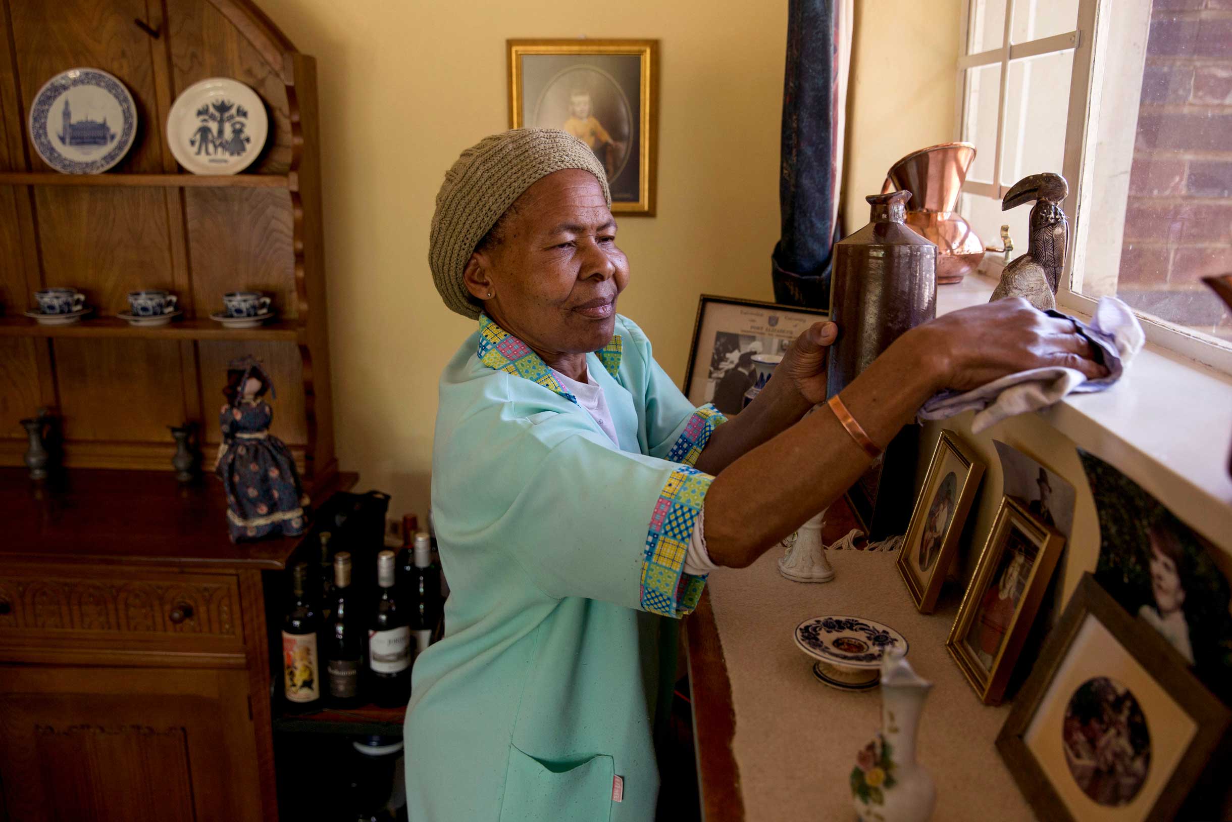 Anna Nkobele, a domestic worker in a blue uniform, dusts a shelf in a home in Johannesburg.