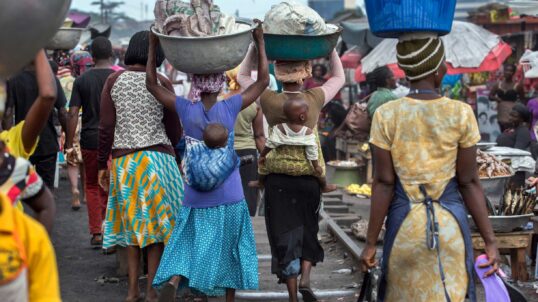 Women carrying goods on their heads walk through a busy market in Ghana.