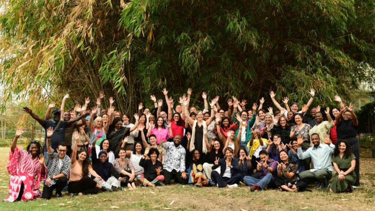 The WIEGO Team poses together outdoors during a team retreat in Nairobi, Kenya, smiling and raising their hands in celebration