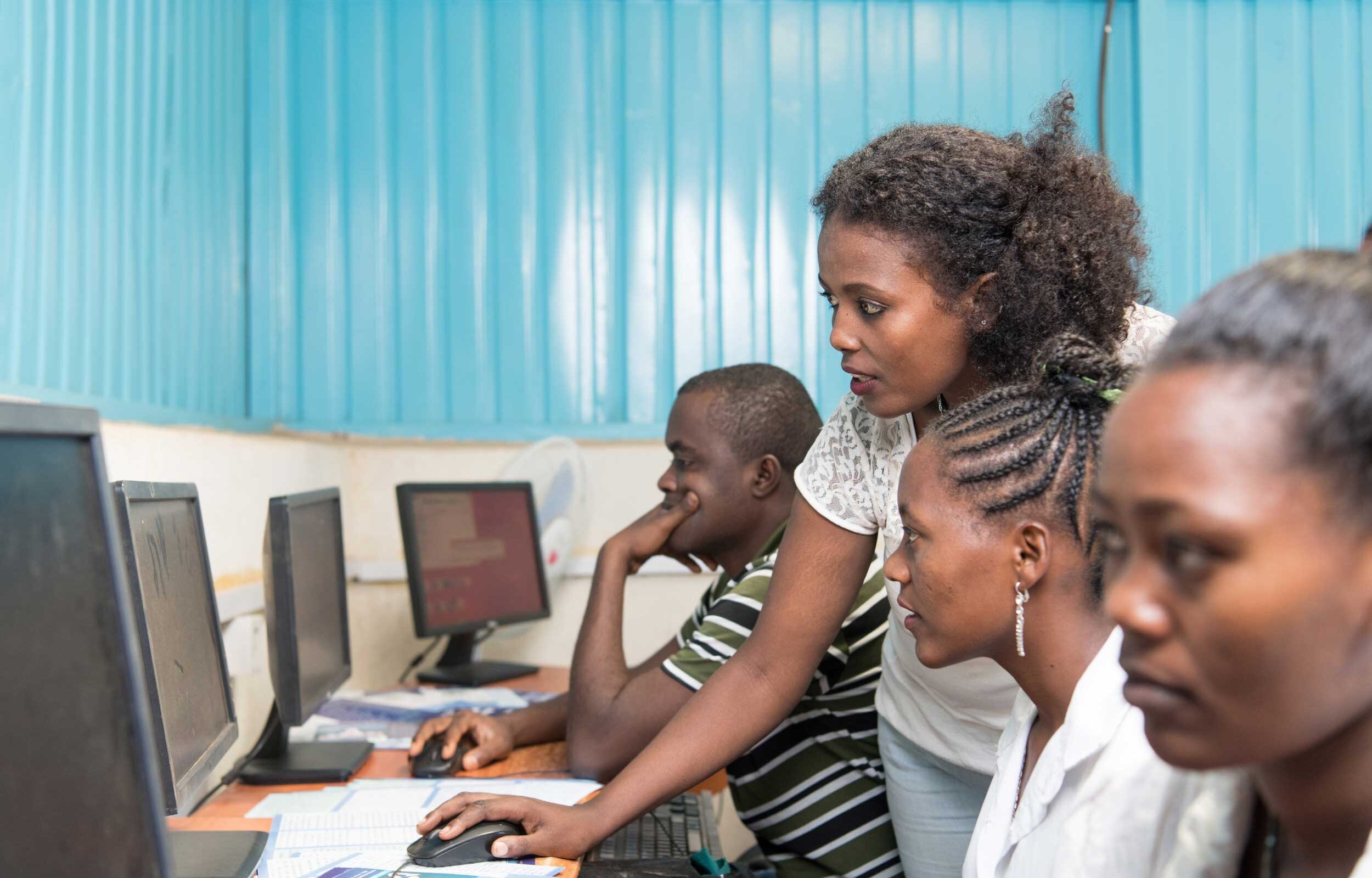 A young woman assisting three students as they work on desktop computers in a training center.