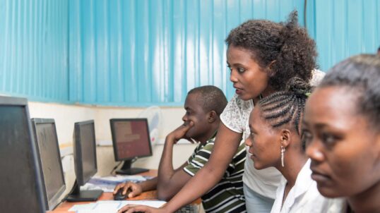 A young woman assisting three students as they work on desktop computers in a training center.