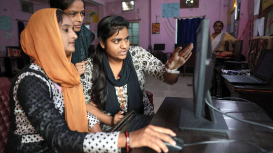 Narmada Ben, an informal worker trained by SEWA and MHT, collaborates with colleagues on a computer as part of her work conducting surveys in her neighborhood slum.