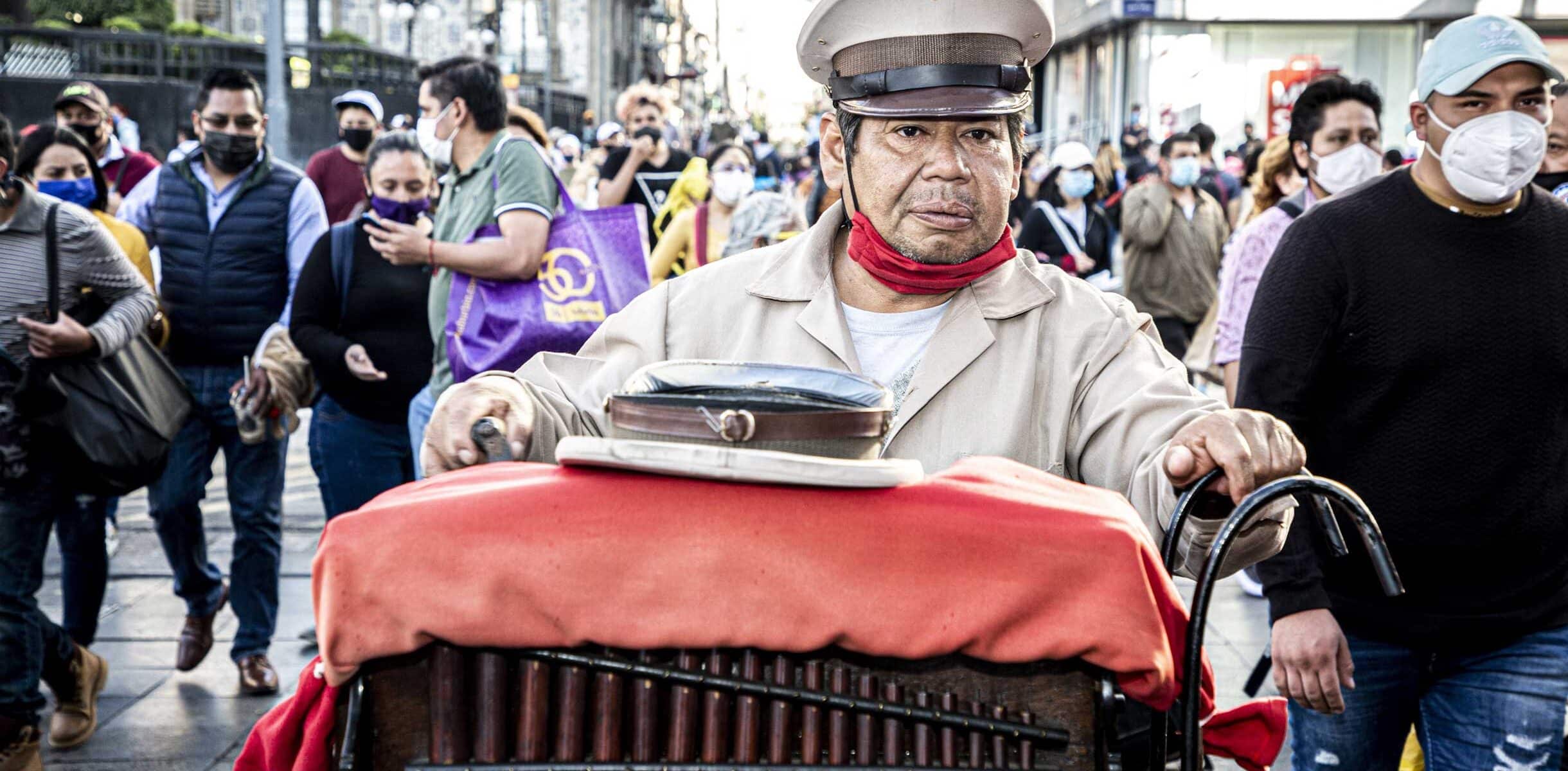 A street organ player wearing a beige uniform and hat, pushing his organ through a busy street in Mexico City, surrounded by pedestrians.