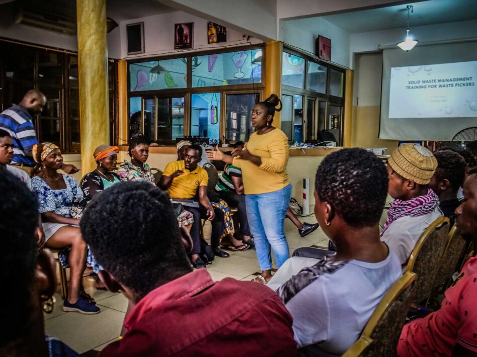 Dorcas Ansah of WIEGO leads a training session on solid waste management with waste pickers in Accra, Ghana, facilitating a discussion with participants. A presentation on waste management is projected in the background.