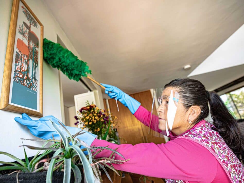 Raquel Martinez, a domestic worker in gloves and a face shield, dusts a framed picture in a home.