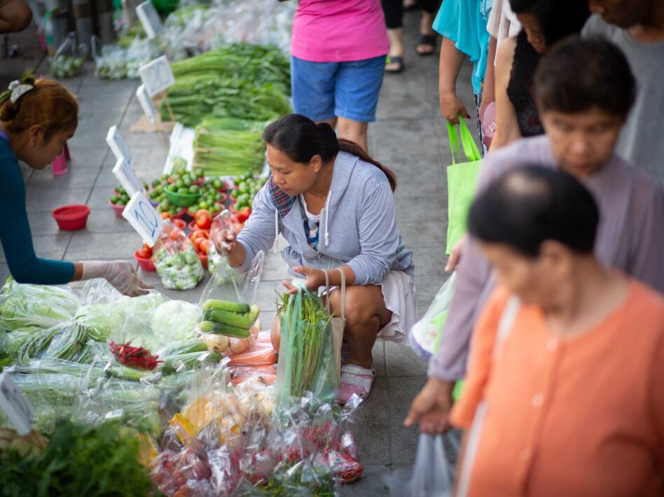 A customer selects fresh vegetables at a vendor’s stall in one of Bangkok’s bustling markets, where vendors and shoppers interact amid a variety of colorful produce.