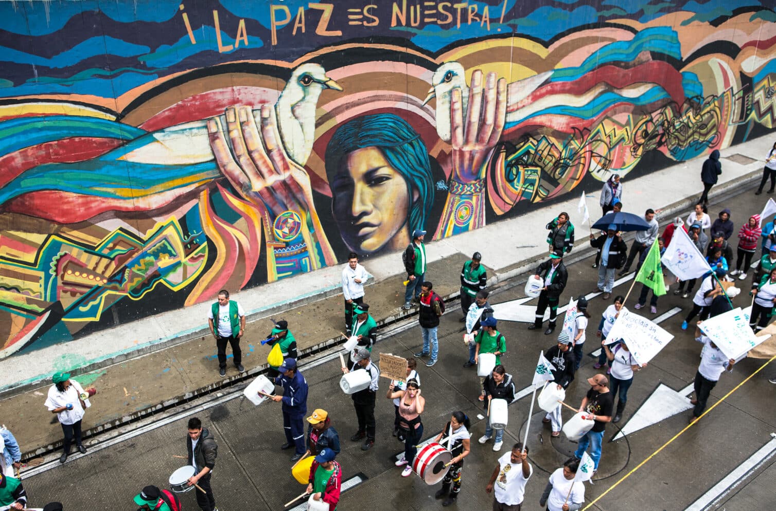 Waste pickers march in Bogotá under a colorful mural with protest signs and drums.