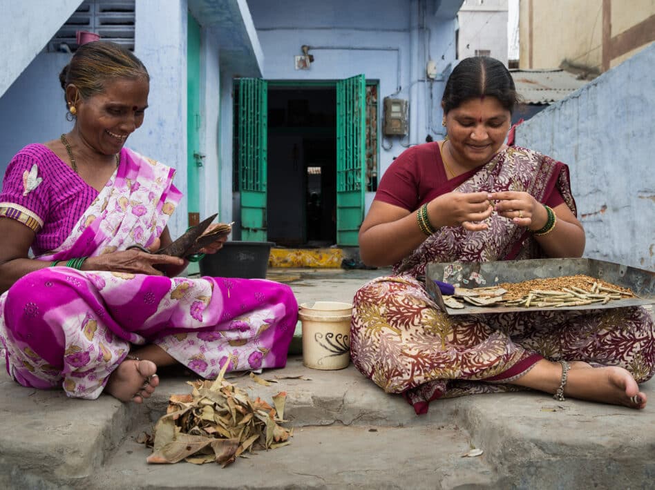 Two Indian women in sarees hand-roll bidis outside their home.