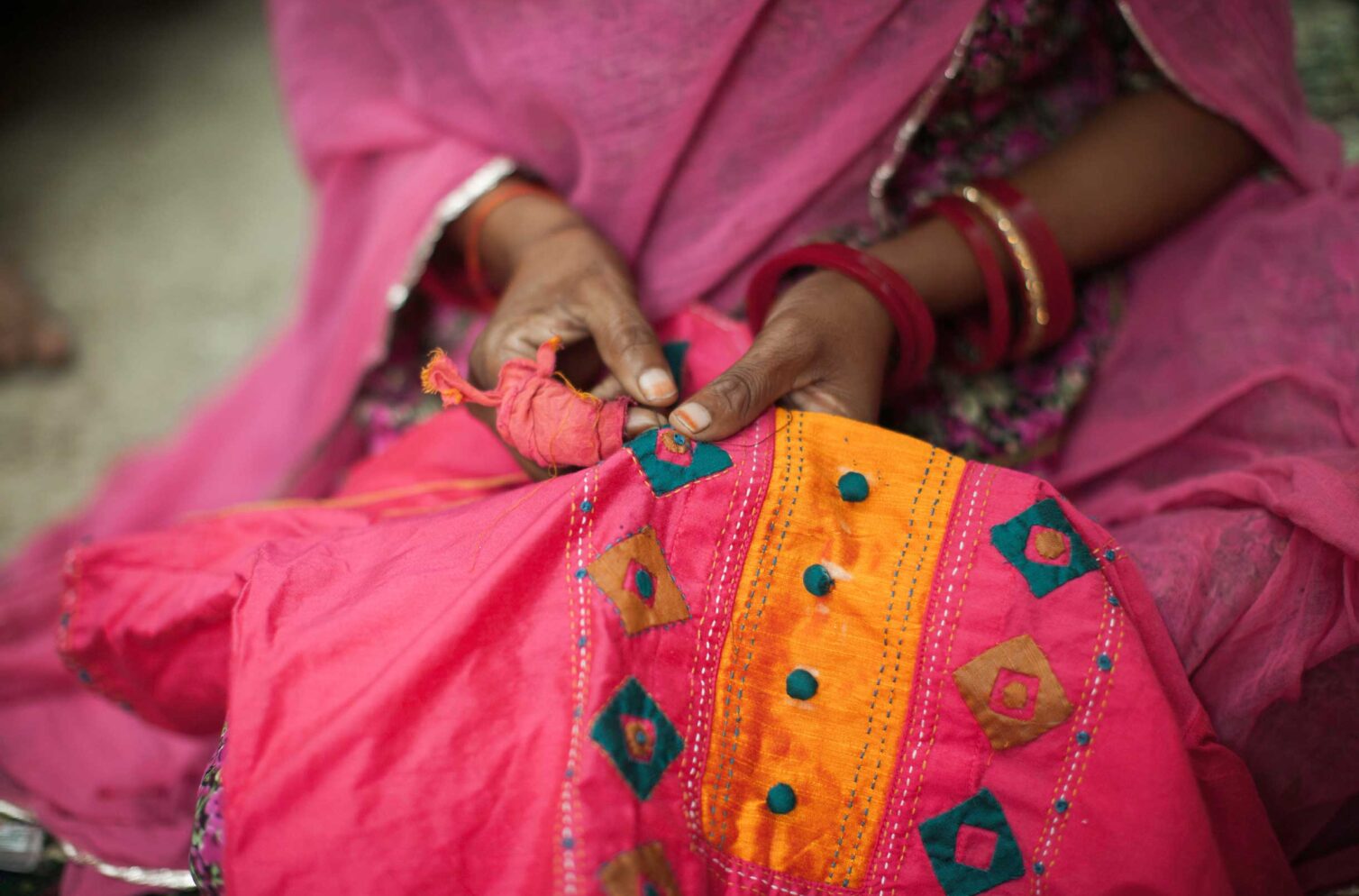 Close-up of a woman’s hands embroidering pink and orange fabric with intricate patterns.