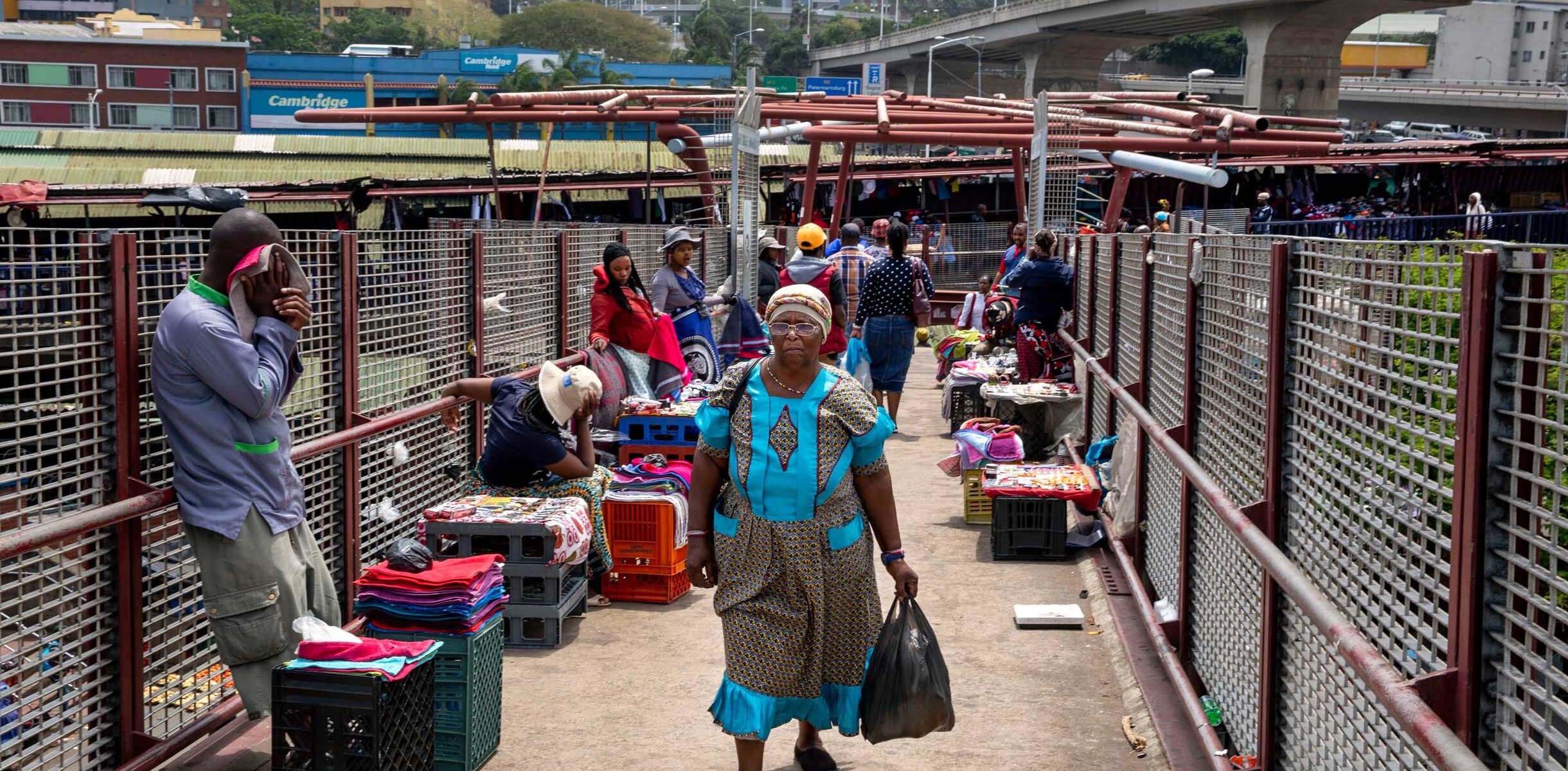 A woman walks through a market bridge in Durban, with vendors selling colorful textiles.